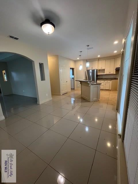 kitchen featuring stainless steel fridge, light tile patterned floors, a center island with sink, white cabinetry, and hanging light fixtures