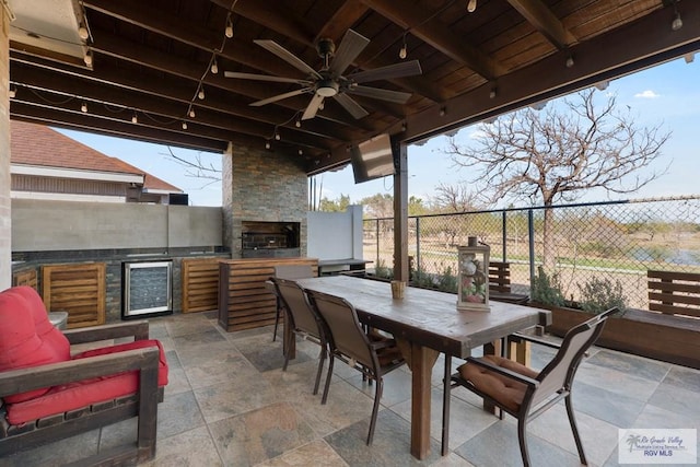 view of patio with beverage cooler, an outdoor stone fireplace, fence, a ceiling fan, and outdoor dining space