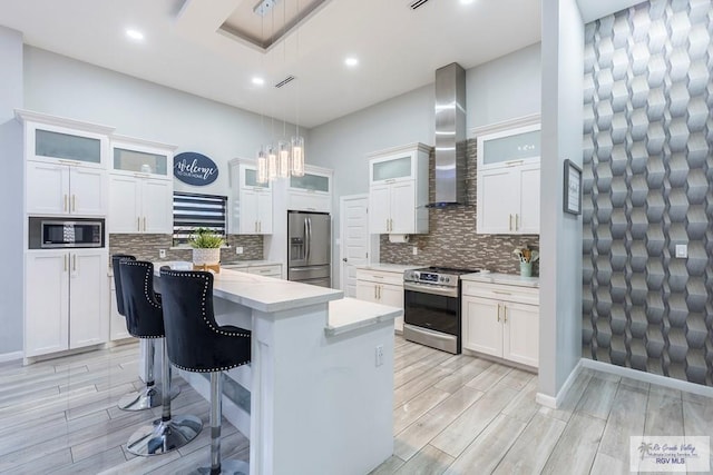 kitchen with wall chimney range hood, hanging light fixtures, appliances with stainless steel finishes, a kitchen island, and white cabinetry