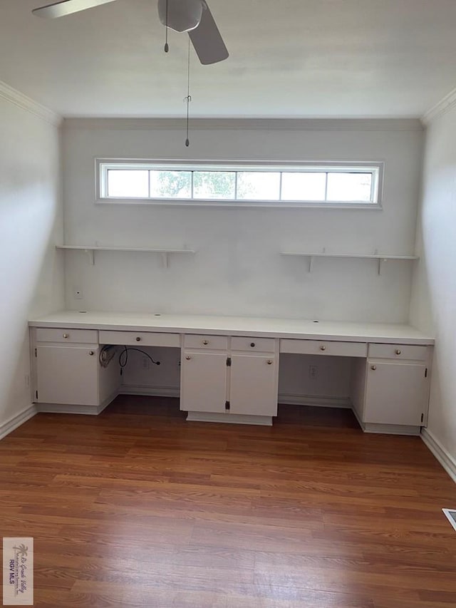 unfurnished office featuring baseboards, ornamental molding, built in desk, a ceiling fan, and dark wood-style flooring