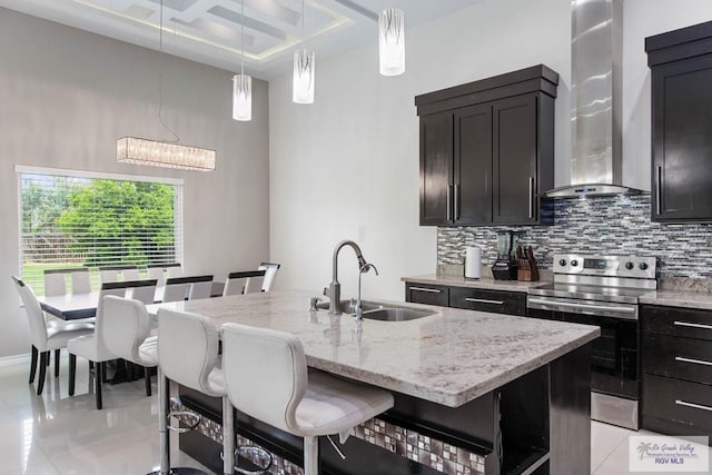 kitchen with electric range, sink, wall chimney exhaust hood, a kitchen breakfast bar, and an island with sink