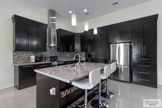 kitchen featuring a kitchen island with sink, wall chimney range hood, sink, decorative light fixtures, and stainless steel appliances