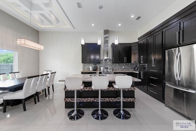 kitchen featuring light stone countertops, appliances with stainless steel finishes, wall chimney range hood, a center island with sink, and a breakfast bar area