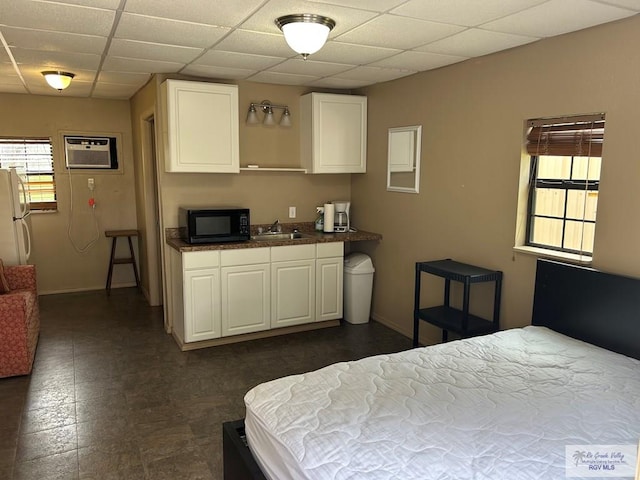 bedroom featuring a wall unit AC, baseboards, a drop ceiling, freestanding refrigerator, and a sink