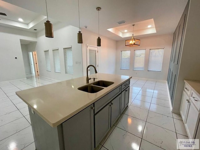 kitchen featuring gray cabinetry, a raised ceiling, sink, pendant lighting, and an island with sink