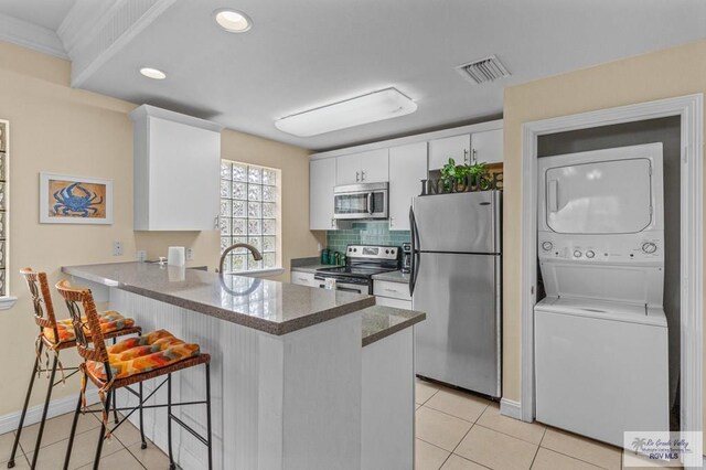 106 Atol St Unit 7, South Padre Island, TX 78597 - kitchen featuring appliances with stainless steel finishes, stacked washing machine and dryer, light tile patterned flooring, a peninsula, and white cabinets