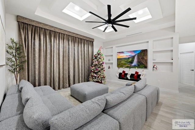 living room featuring a skylight, light wood-type flooring, built in features, ceiling fan, and a tray ceiling