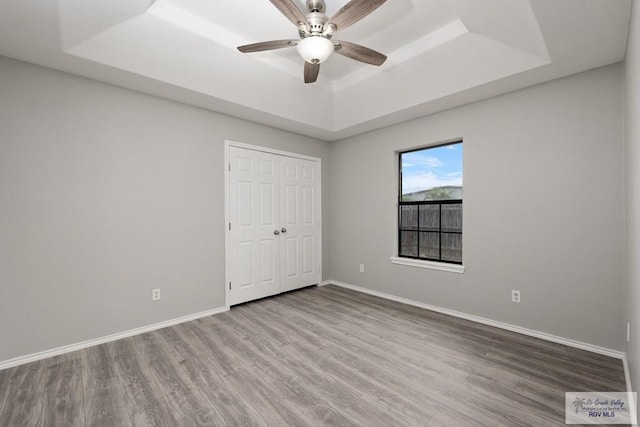 unfurnished bedroom with a raised ceiling, a closet, a ceiling fan, wood finished floors, and baseboards