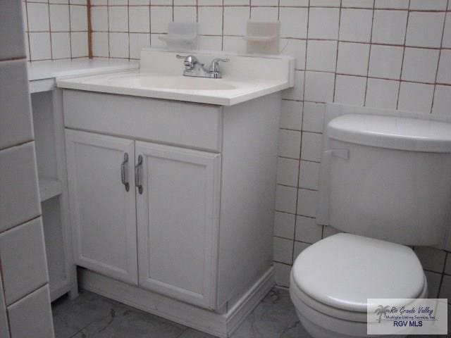 half bath with toilet, marble finish floor, tile walls, and vanity