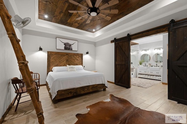 bedroom with ensuite bathroom, a tray ceiling, ceiling fan, a barn door, and light hardwood / wood-style flooring