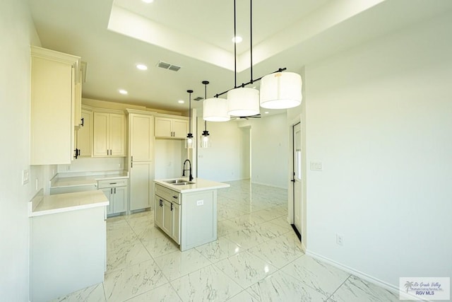 kitchen with sink, a center island with sink, a tray ceiling, pendant lighting, and white cabinets