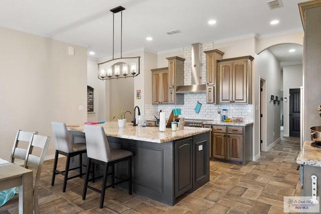 kitchen featuring backsplash, light stone counters, ornamental molding, wall chimney exhaust hood, and a kitchen island with sink