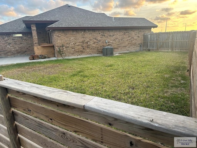 view of side of home with central AC unit, fence, a shingled roof, a yard, and brick siding