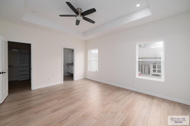 unfurnished bedroom featuring light wood-type flooring, ensuite bathroom, a spacious closet, and a tray ceiling