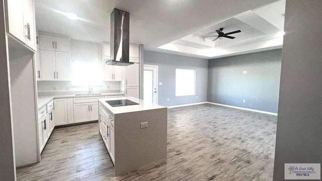 kitchen featuring wall chimney exhaust hood, a kitchen island, a raised ceiling, black electric cooktop, and white cabinets