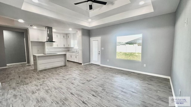 kitchen with light wood-type flooring, wall chimney exhaust hood, a tray ceiling, white cabinets, and a kitchen island