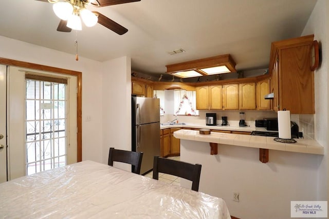 kitchen with a kitchen breakfast bar, stainless steel fridge, kitchen peninsula, tile countertops, and ceiling fan