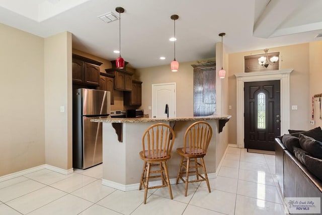 kitchen with a breakfast bar, decorative light fixtures, dark brown cabinets, light stone counters, and stainless steel refrigerator