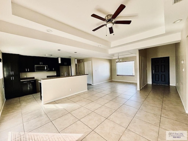 kitchen featuring stainless steel appliances, a tray ceiling, a kitchen island with sink, light tile patterned flooring, and ceiling fan with notable chandelier