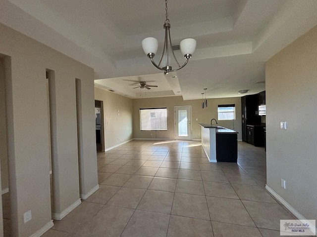 kitchen with a tray ceiling, a kitchen island with sink, light tile patterned floors, and ceiling fan with notable chandelier