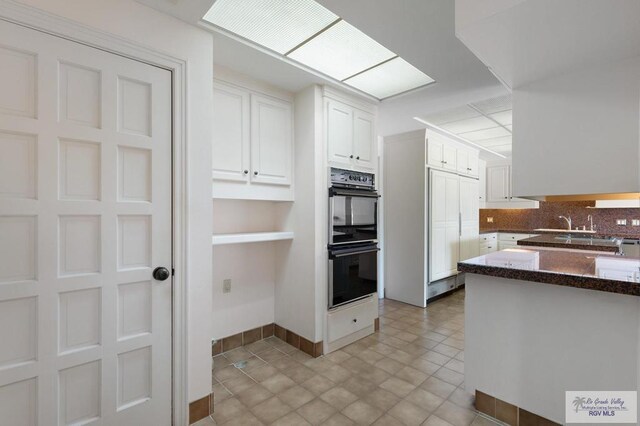 kitchen featuring decorative backsplash, dark stone counters, double oven, sink, and white cabinets