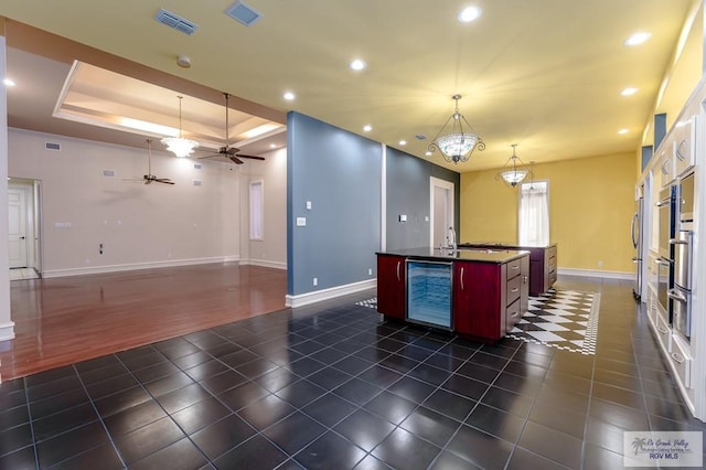 kitchen featuring beverage cooler, a raised ceiling, dark hardwood / wood-style flooring, decorative light fixtures, and a center island with sink