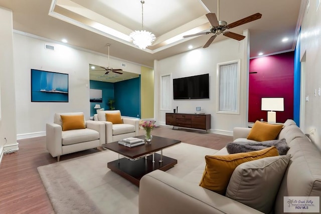 living room with wood-type flooring, ceiling fan with notable chandelier, a tray ceiling, and crown molding