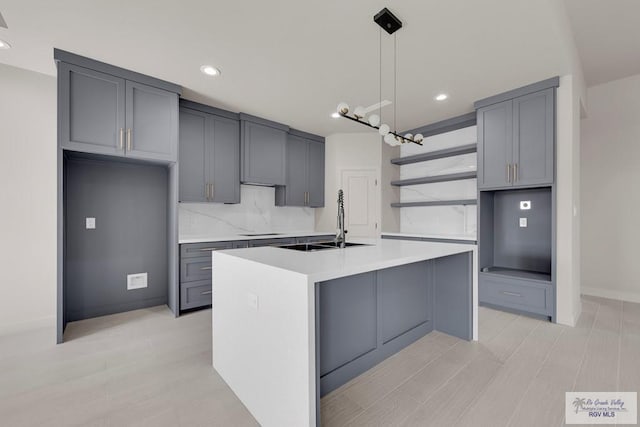 kitchen with gray cabinetry, an island with sink, a sink, open shelves, and light countertops