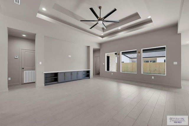 unfurnished living room with visible vents, ceiling fan, baseboards, a tray ceiling, and light wood-style floors