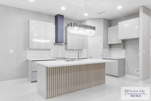 kitchen with white cabinetry, sink, wall chimney exhaust hood, hanging light fixtures, and a center island with sink