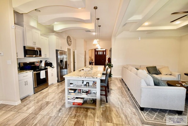 kitchen with appliances with stainless steel finishes, white cabinetry, hanging light fixtures, light stone countertops, and an island with sink