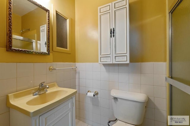 full bathroom featuring a shower with shower door, a wainscoted wall, toilet, vanity, and tile walls