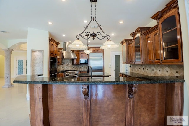 kitchen with kitchen peninsula, decorative columns, premium appliances, wall chimney range hood, and hanging light fixtures