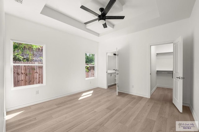 unfurnished bedroom with a tray ceiling, light wood-style flooring, a spacious closet, ceiling fan, and baseboards