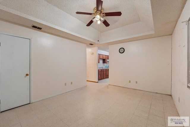 empty room with a textured ceiling, ceiling fan, visible vents, light floors, and a tray ceiling