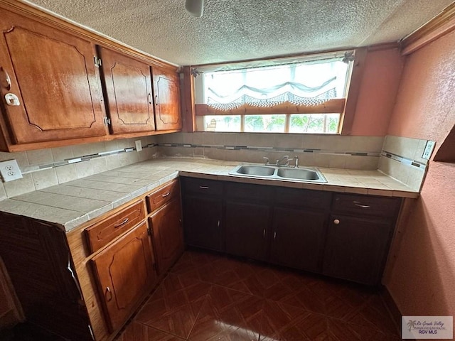 1022 E Monroe Ave, Harlingen, TX 78550 - kitchen with a sink, a textured ceiling, decorative backsplash, brown cabinetry, and tile counters