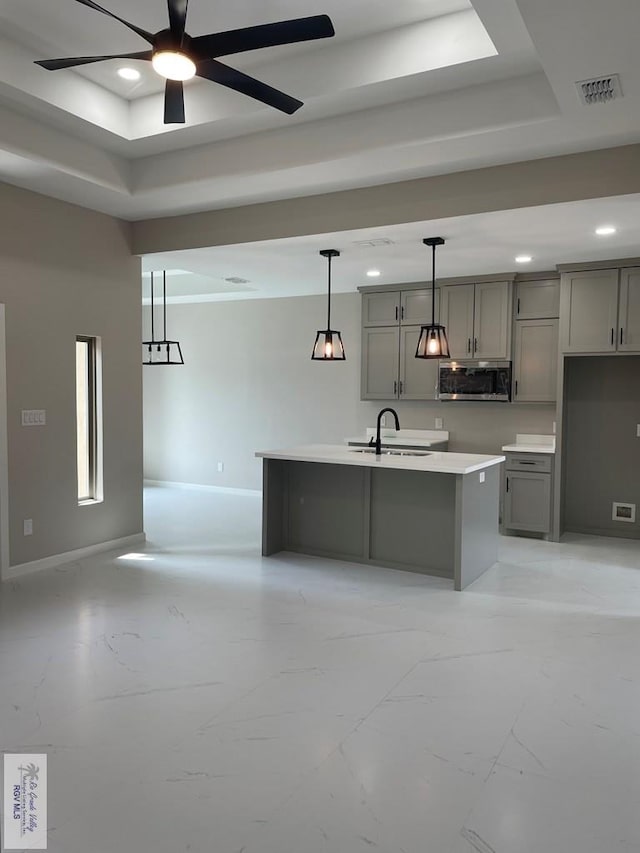 kitchen featuring stainless steel microwave, a kitchen island with sink, a tray ceiling, gray cabinets, and a sink