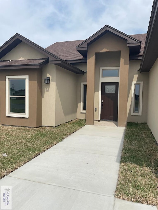 doorway to property featuring a shingled roof, a lawn, and stucco siding