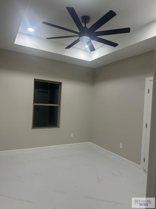 spare room with a tray ceiling, marble finish floor, ceiling fan, and baseboards