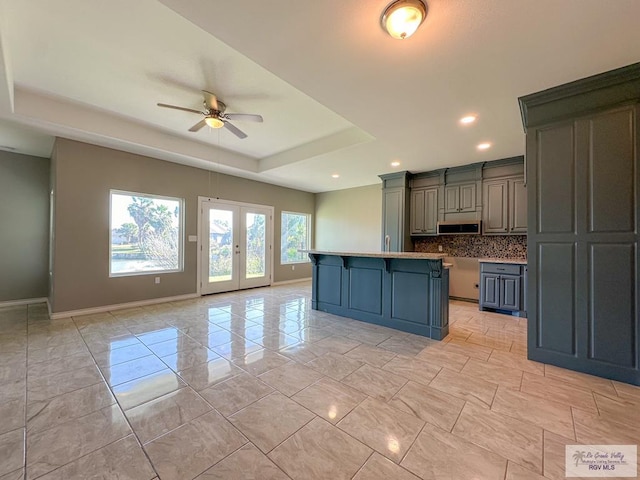 kitchen with french doors, a tray ceiling, an island with sink, ceiling fan, and decorative backsplash