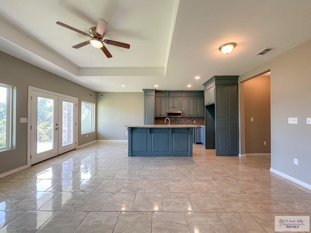 kitchen with french doors, a tray ceiling, ceiling fan, a kitchen island with sink, and backsplash