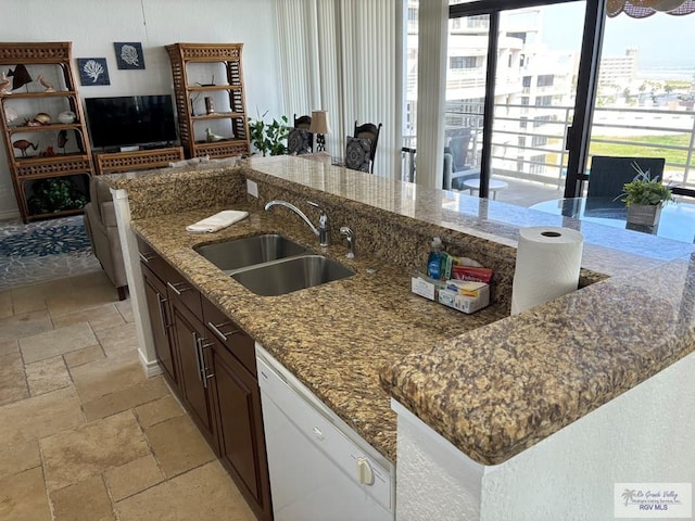 kitchen featuring white dishwasher, stone tile flooring, a sink, dark stone countertops, and a center island with sink
