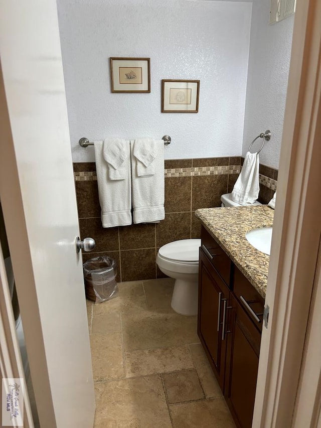 half bathroom featuring tile walls, stone tile floors, toilet, wainscoting, and vanity