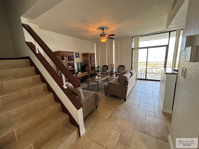 living room featuring a ceiling fan, a wall of windows, stairway, and stone tile flooring
