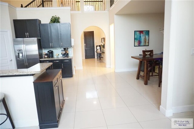 kitchen with backsplash, stainless steel refrigerator with ice dispenser, light stone countertops, light tile patterned floors, and a towering ceiling