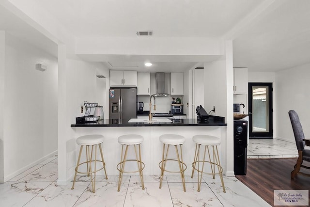 kitchen with kitchen peninsula, stainless steel refrigerator with ice dispenser, a kitchen bar, wall chimney range hood, and white cabinetry