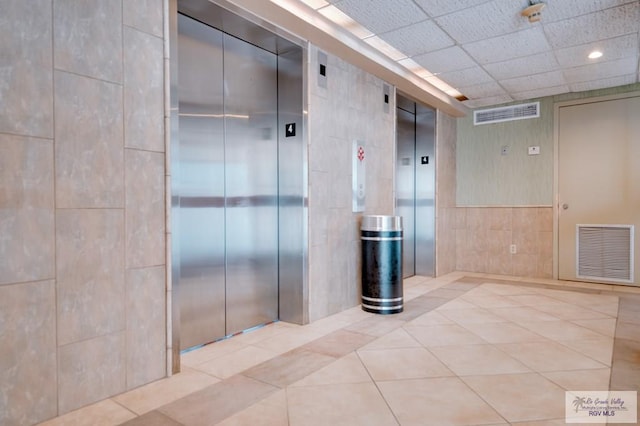 hallway featuring a drop ceiling, light tile patterned floors, tile walls, and elevator