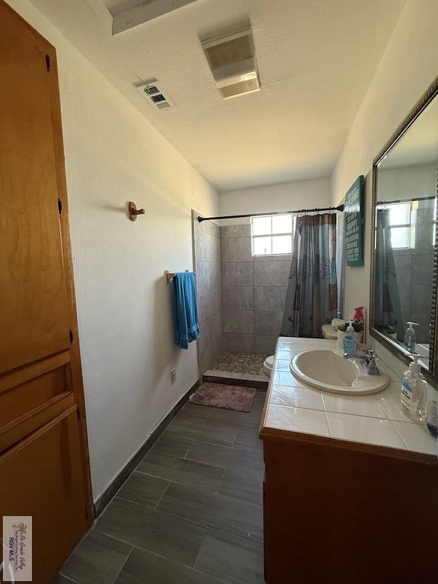 bathroom featuring visible vents, baseboards, toilet, tiled shower, and vanity
