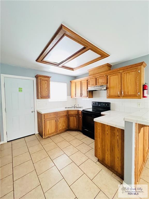 kitchen with tile countertops, brown cabinetry, black range with electric cooktop, under cabinet range hood, and backsplash