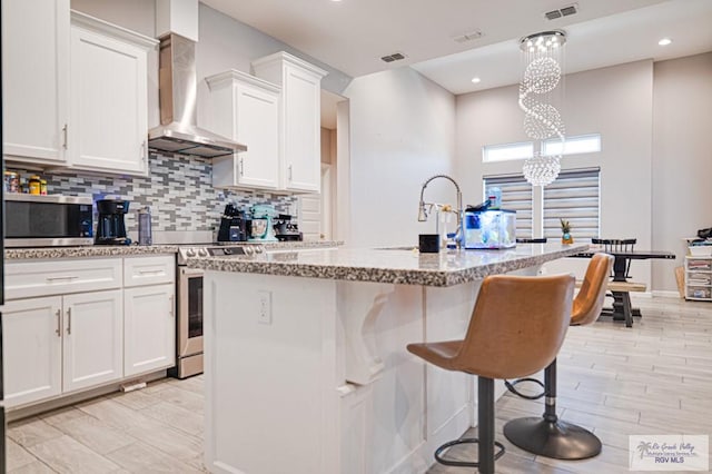 kitchen with an island with sink, wall chimney exhaust hood, appliances with stainless steel finishes, decorative light fixtures, and white cabinetry